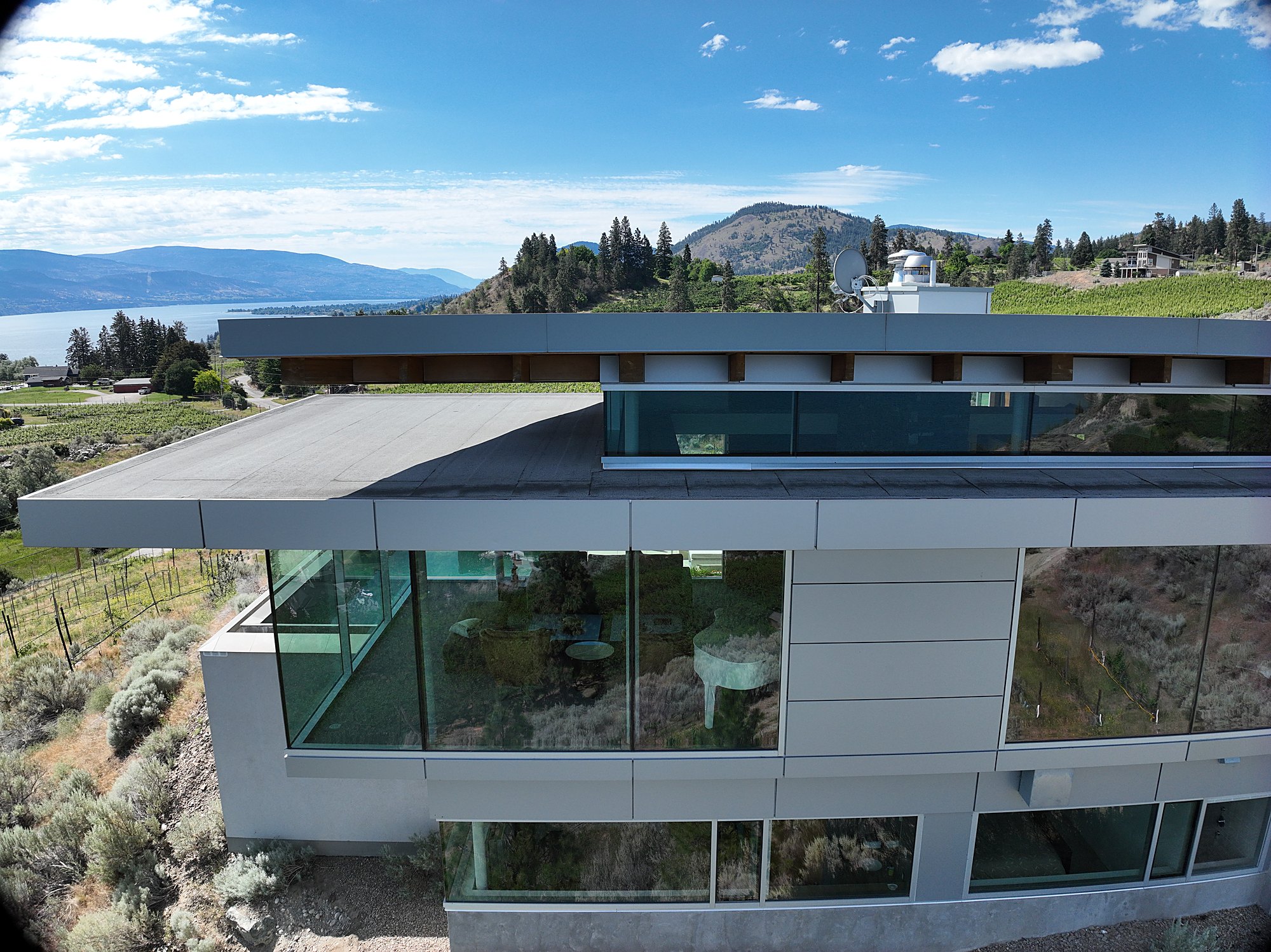 Modern glass house on a hillside above Okanagan Lake, the water and hills behind it.