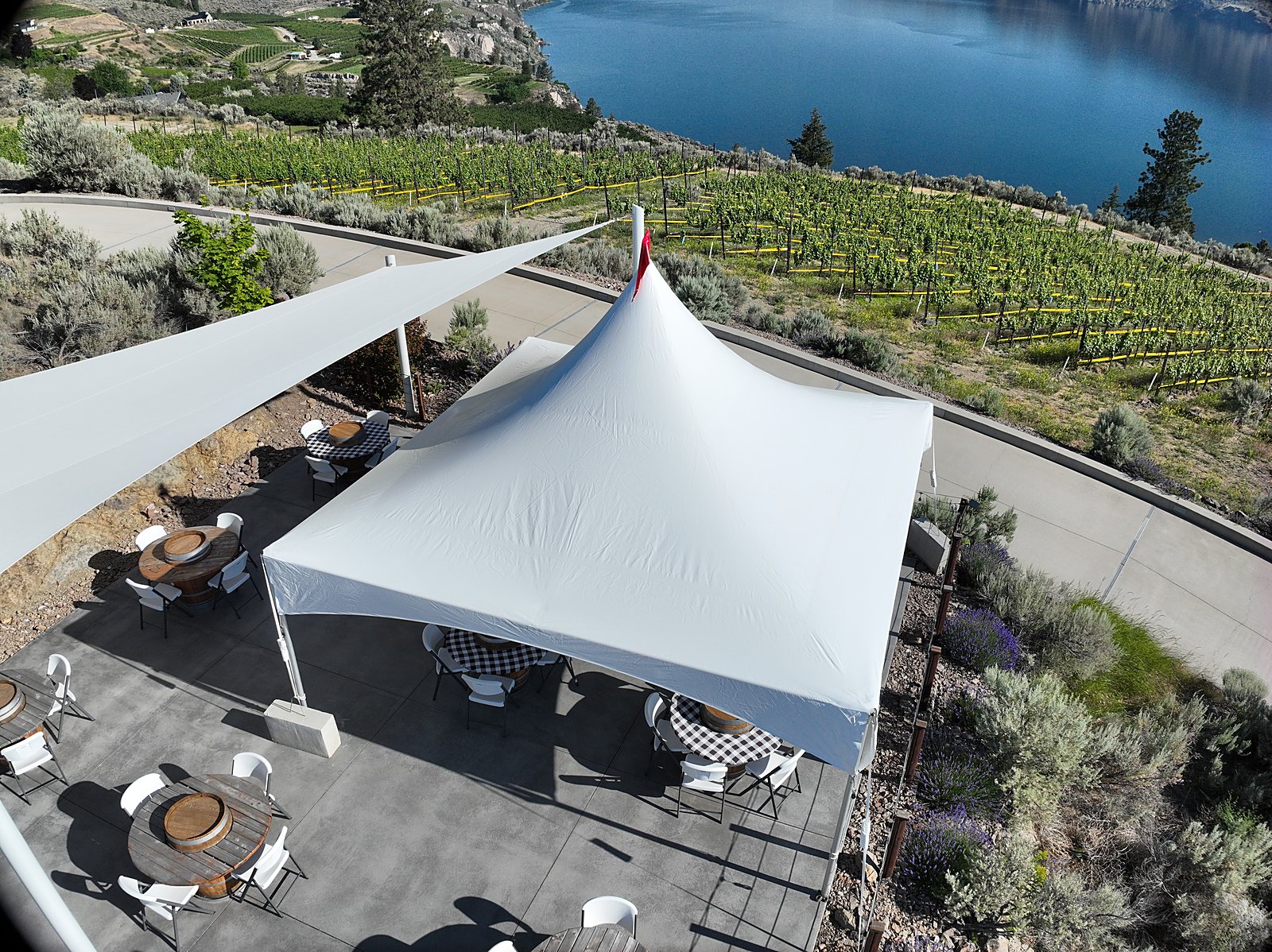 The white-canopied tasting patio of an Okanagan winery from above, barrel tables under the tent and vineyard rows running down to the lake, the kind of hero image a destination winery leads its brand with.